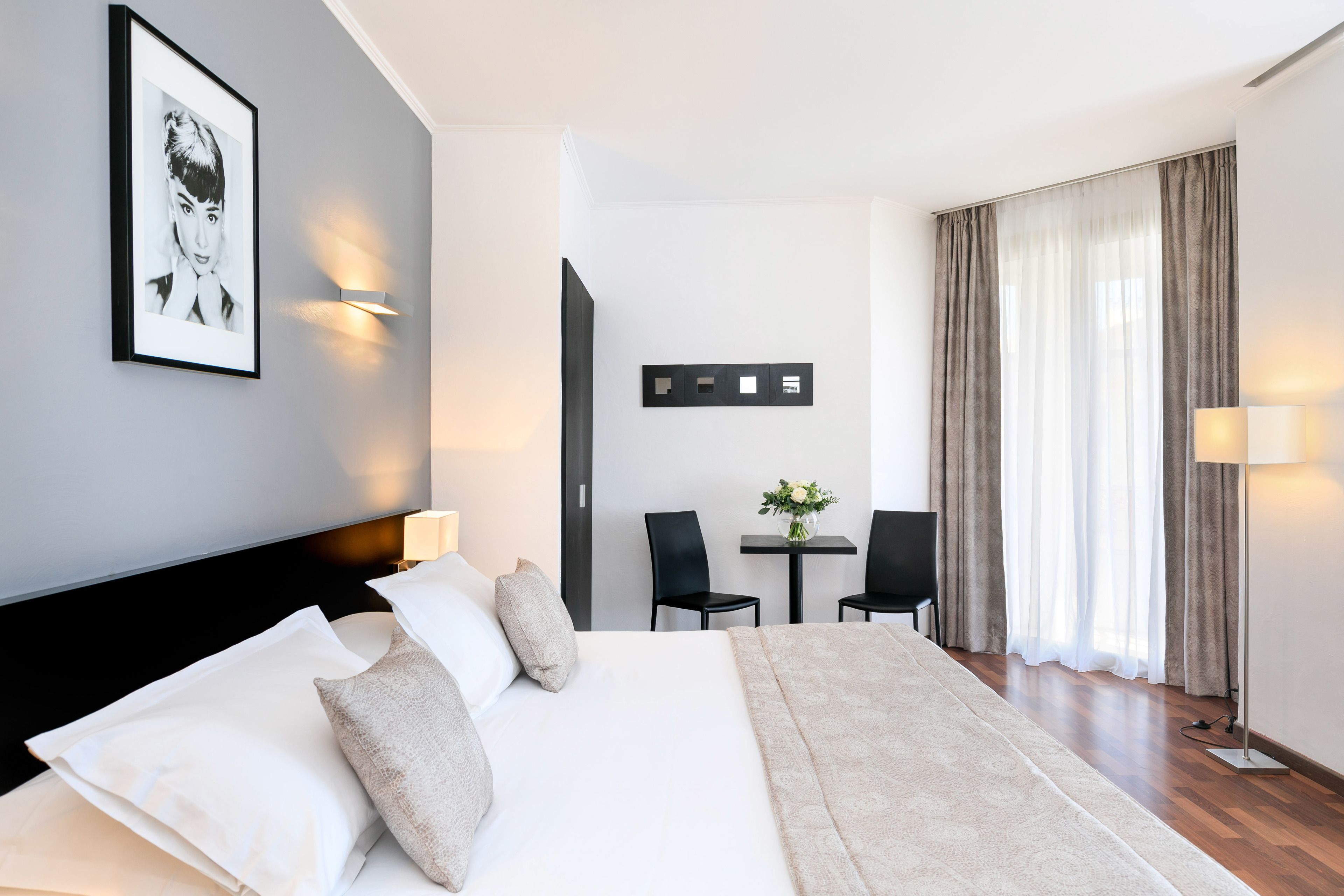 Una habitación de hotel con una cama grande, un escritorio con dos sillas, una mesita con un florero y un ventanal con cortinas.