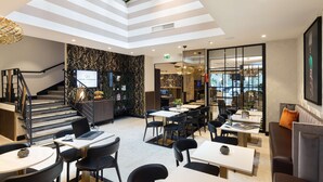 A modern hotel lobby with a high ceiling, a staircase, and a seating area with tables and chairs.