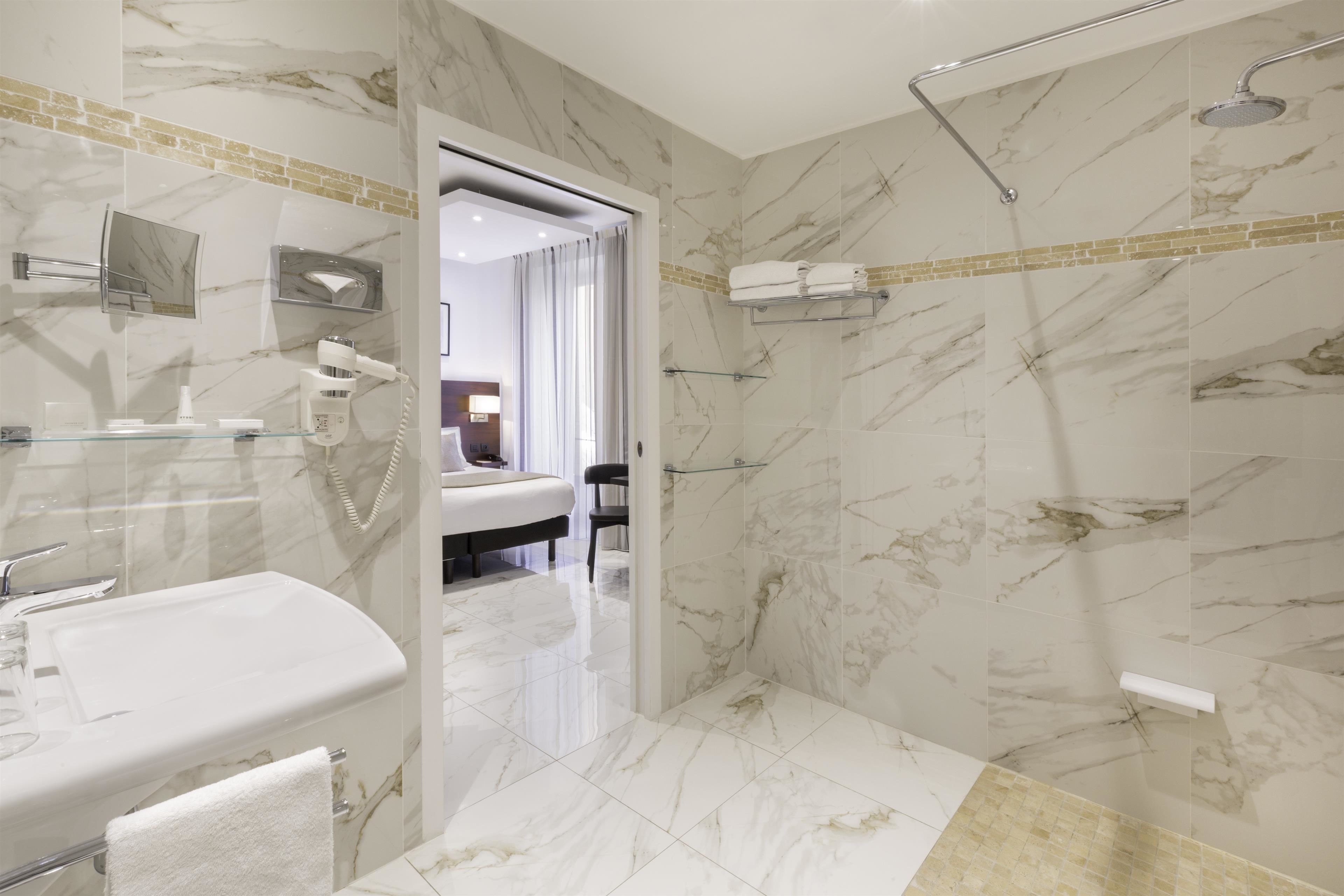 Un baño moderno con ducha de mármol, tocador con recinto de vidrio y una habitación visible al fondo.