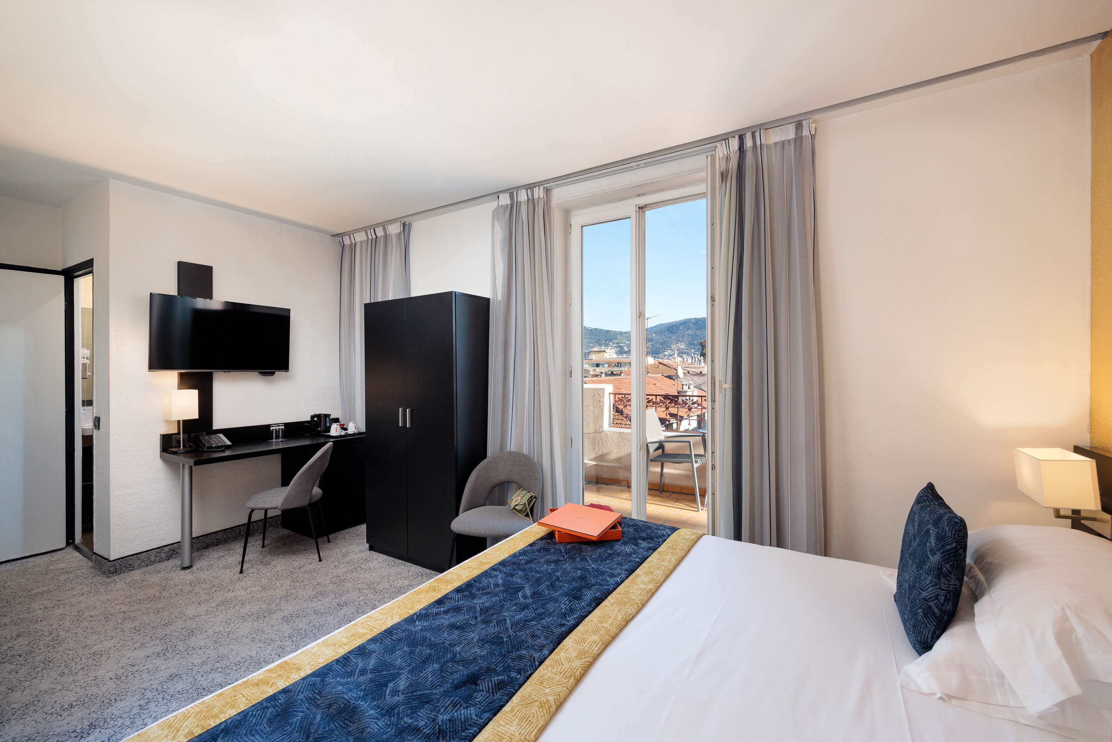 A hotel room with a bed, a desk, a chair, a TV, and a balcony with a view.