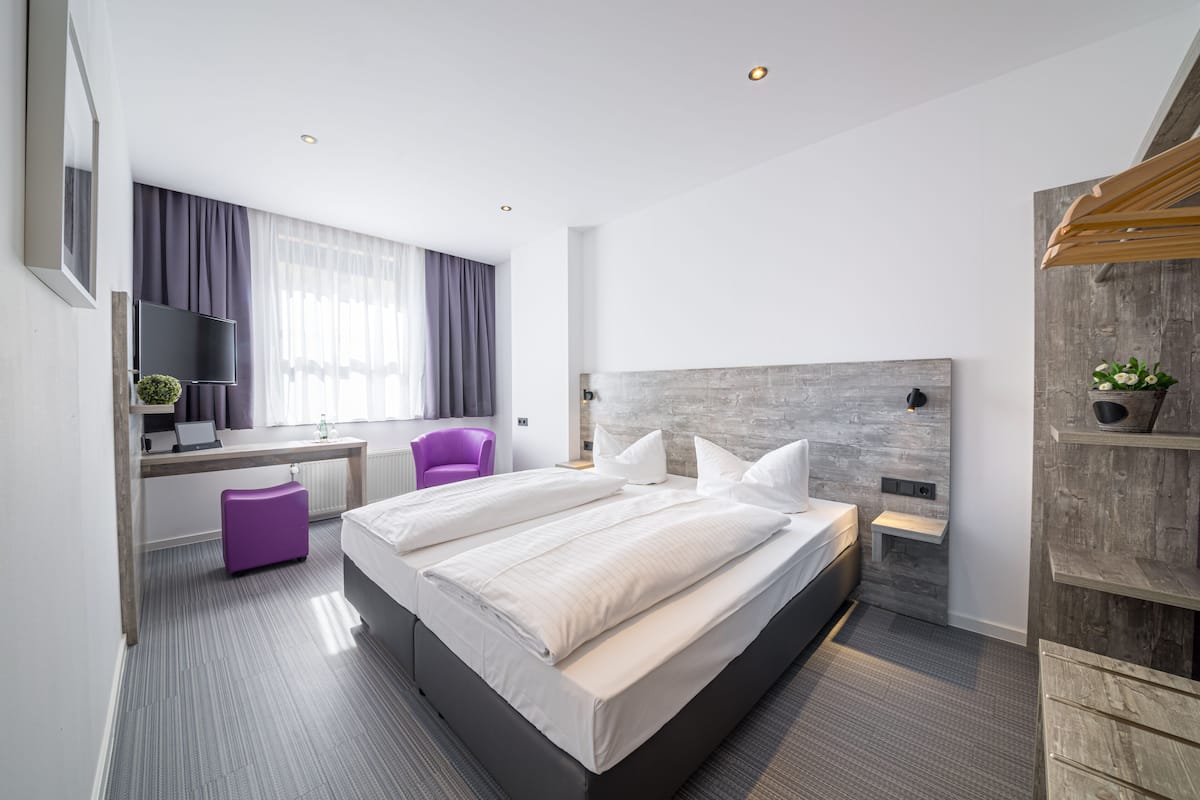 A modern hotel room with a large bed, a desk with a laptop, a purple chair, and a TV.