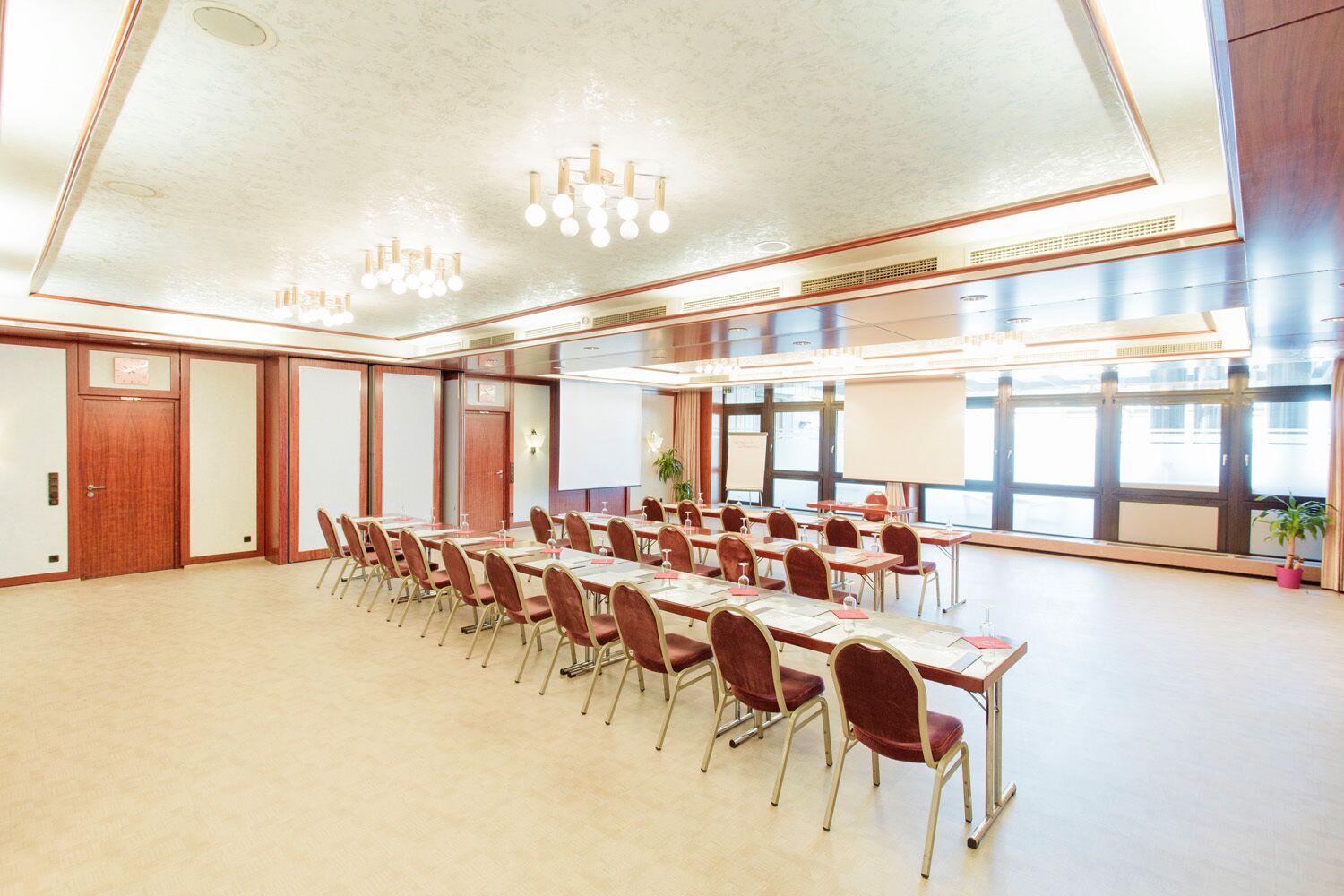 A conference room with multiple tables and chairs, large windows, and a high ceiling with chandeliers.