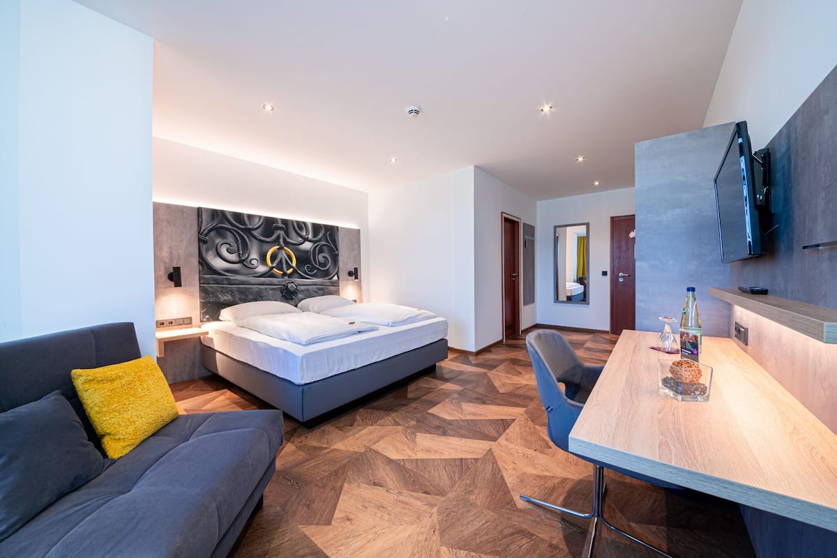 A modern hotel room with a bed, a sofa, a wooden dining table, and a TV mounted on the wall.