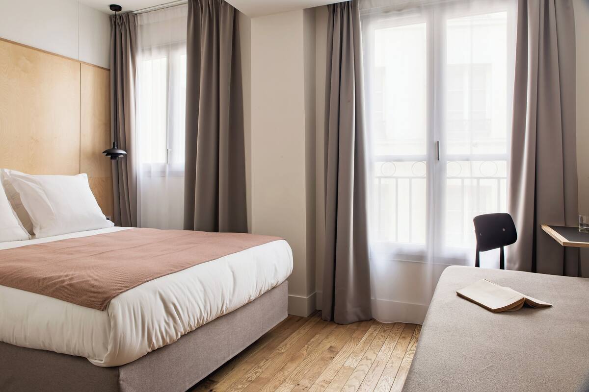 A hotel room with a bed, a chair, a desk, and a large window with curtains.