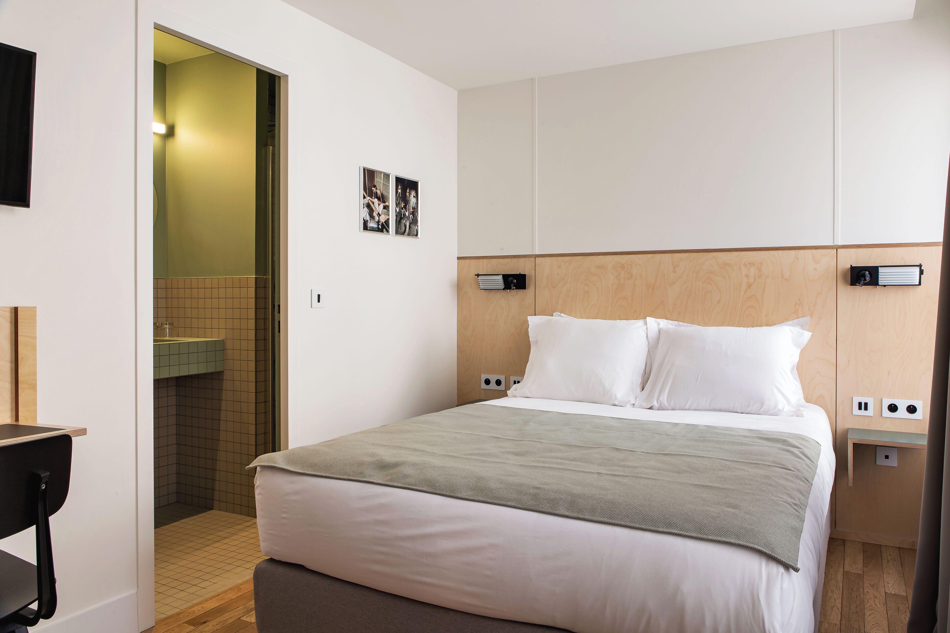 Habitación de hotel ordenada con una cama grande, una mesita de noche, un tocador, cabecera de madera y un baño pequeño con lavamanos y espejo.
