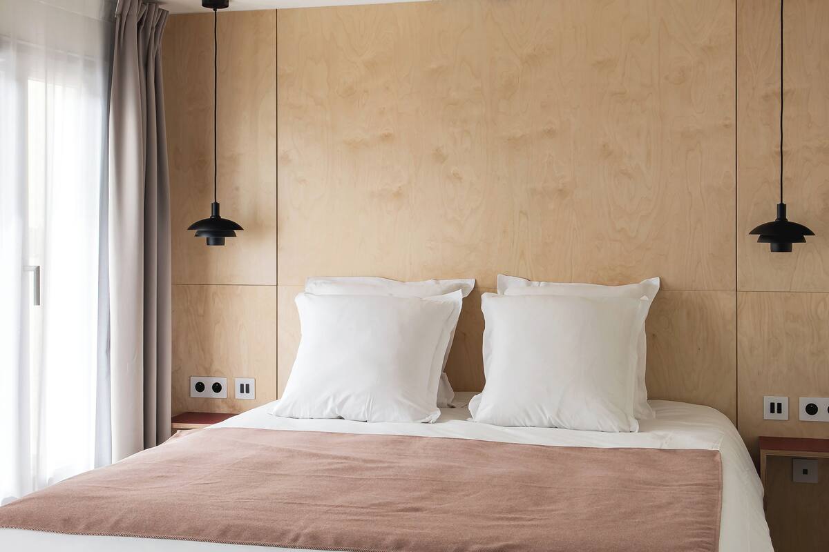 A neatly made bed with a brown throw, wooden headboard, and two pendant lights.