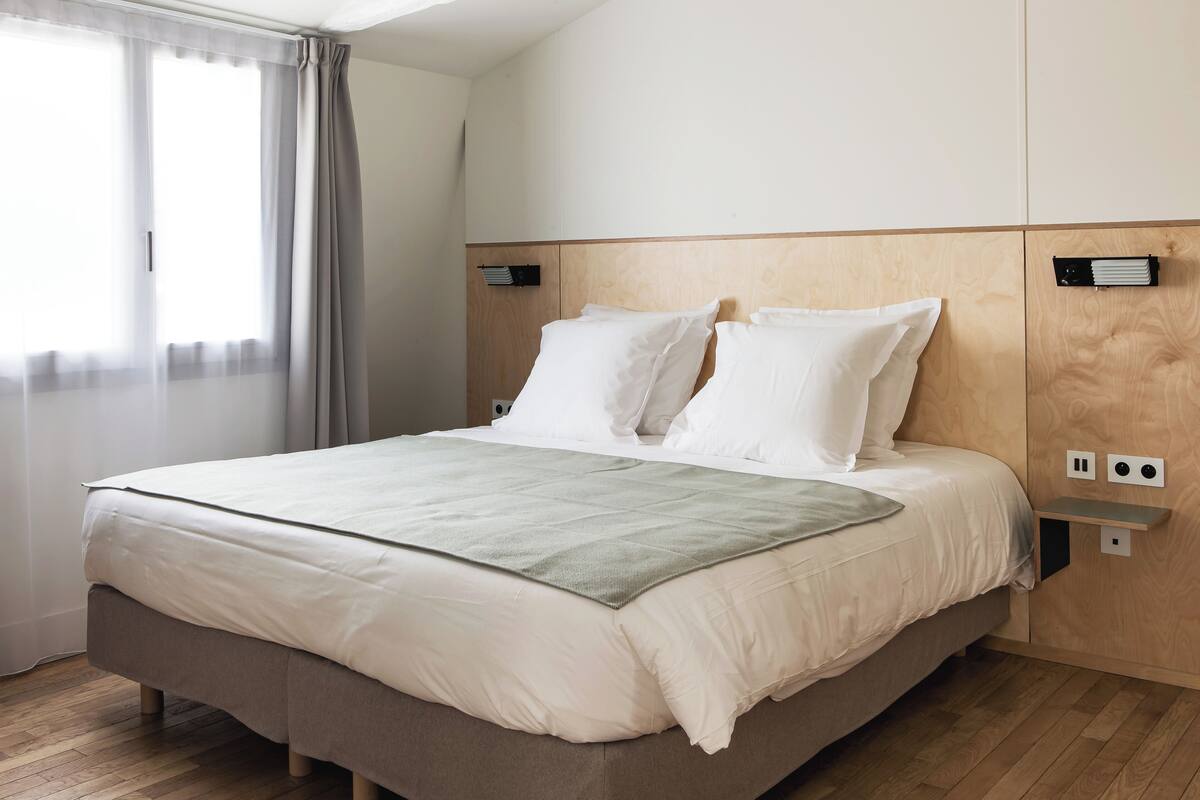 A neatly made bed with a wooden headboard, a bedside table with a power outlet, and a window with sheer curtains.