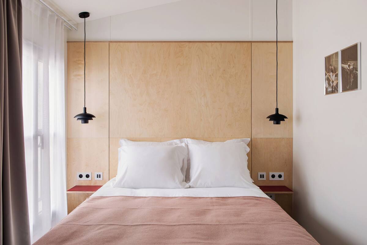 A neatly made bed with white pillows and a pink blanket, wooden headboard, and two pendant lights.
