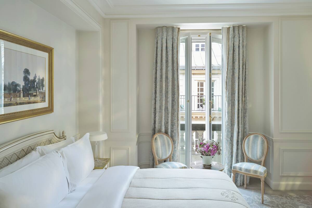 A hotel room with a bed, two chairs, a table with a vase of flowers, and a balcony with curtains.