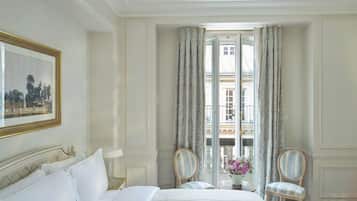 Habitación de hotel con una cama, dos sillas, una mesa con un jarrón de flores y un balcón con cortinas.