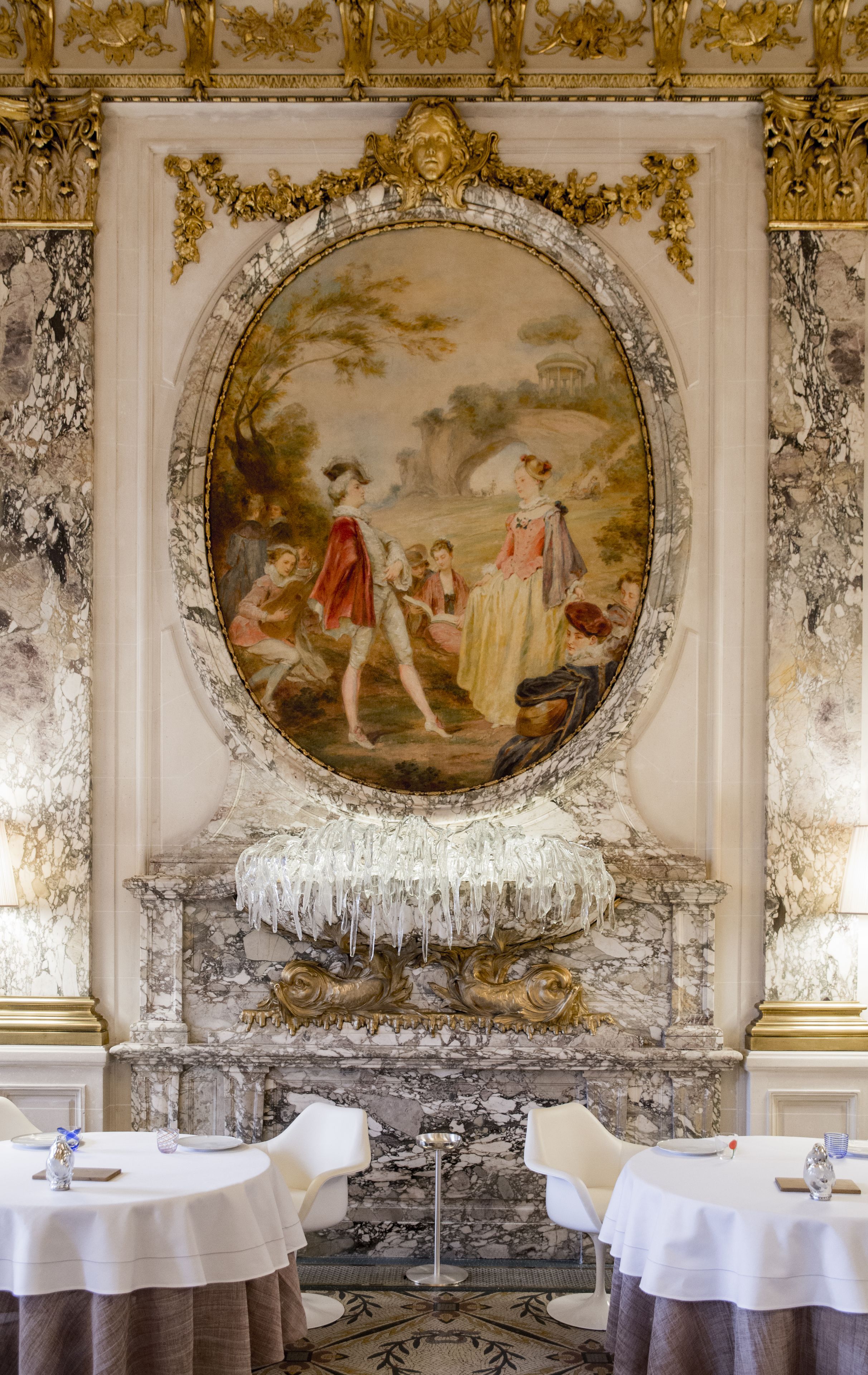 Une vaste pièce agrémentée d’un grand tableau décoratif, de murs en marbre et d’une cheminée surmontée d’un manteau de couleur blanche. Des tables recouvertes de nappes blanches et des chaises sont disposées pour les repas.