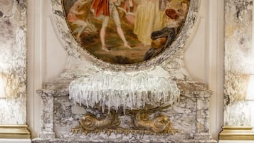 Une vaste pièce agrémentée d’un grand tableau décoratif, de murs en marbre et d’une cheminée surmontée d’un manteau de couleur blanche. Des tables recouvertes de nappes blanches et des chaises sont disposées pour les repas.