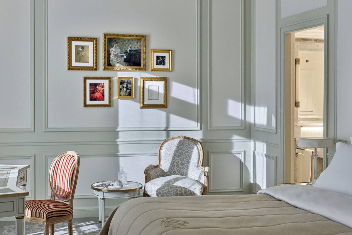 A bedroom with a bed, two chairs, a small table, and a wall with framed pictures.