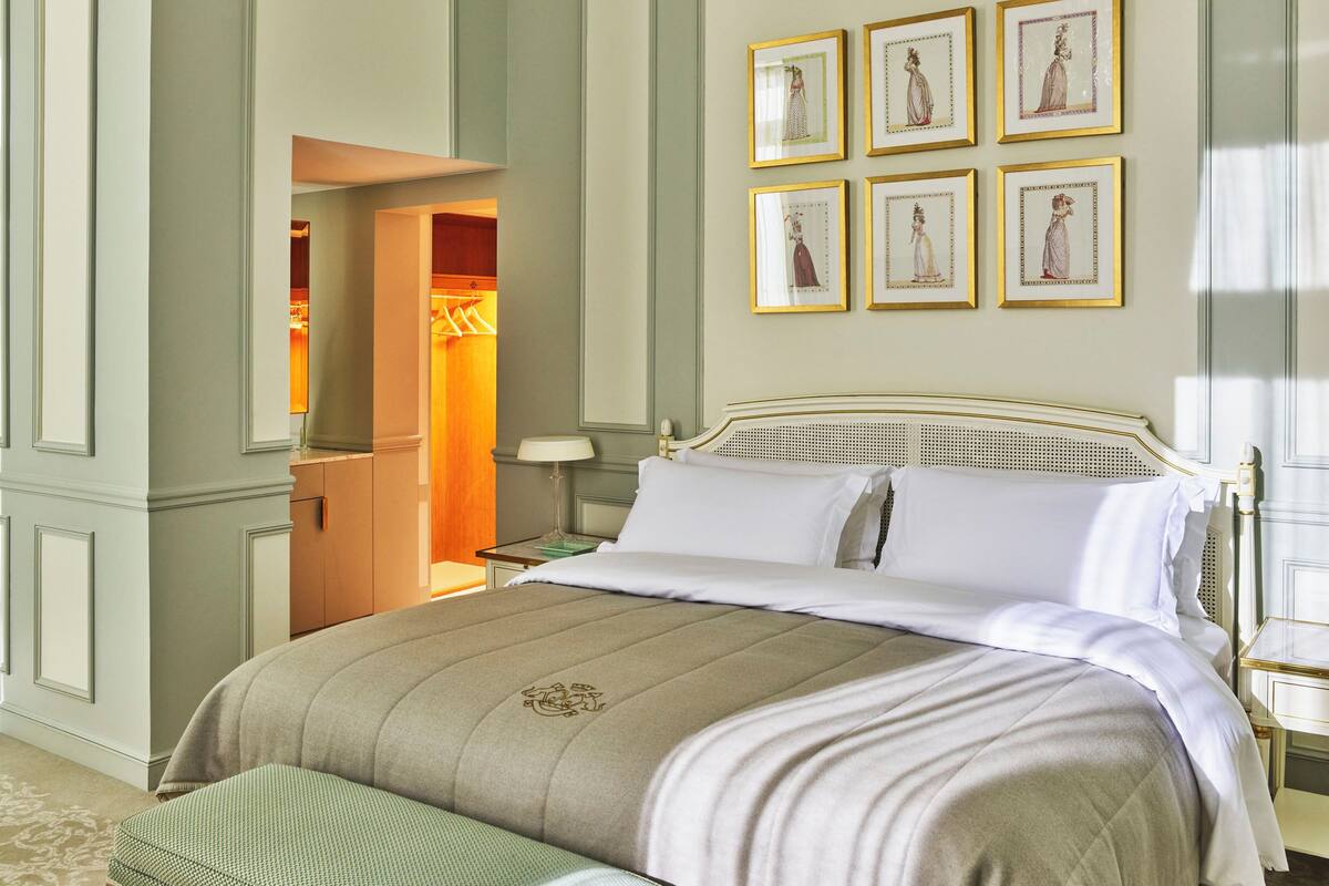 A neatly made bed with a white and beige bedspread, a wooden headboard, and a footboard. There are framed pictures on the wall above the bed.