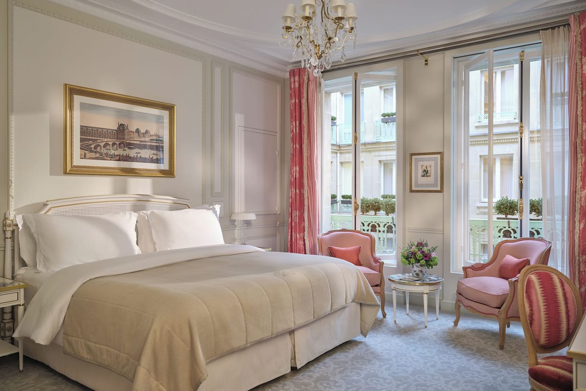A hotel room with a large bed, a chandelier, a painting, and a view of the city through large windows.