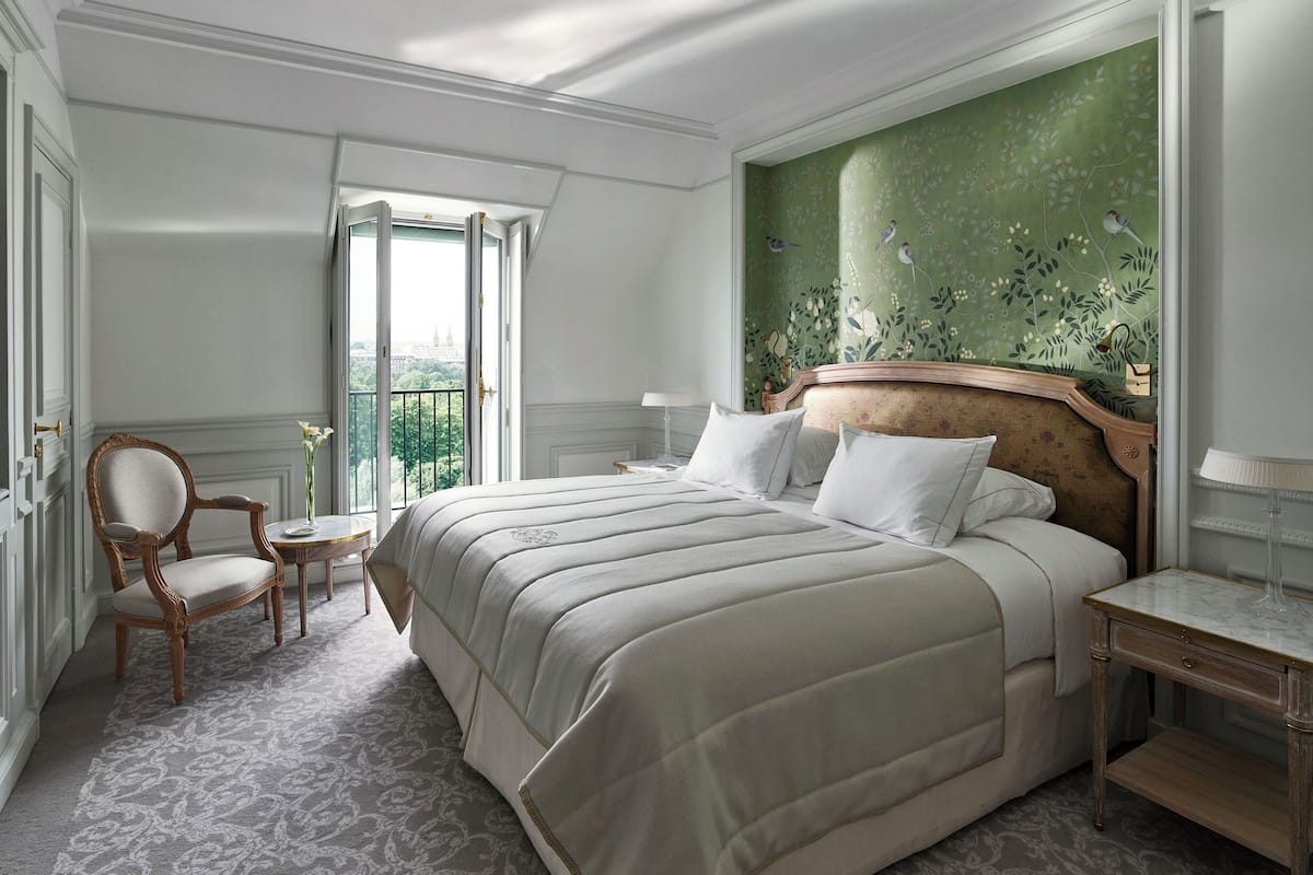 A hotel room with a large bed, a chair, a small table, and a view of the city through a large window.