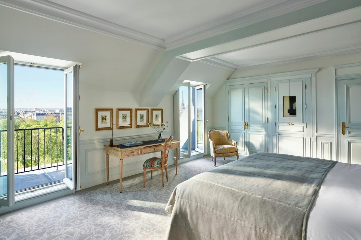 A bedroom with a large bed, a desk with a chair, a wardrobe, and a view of the city through an open door.