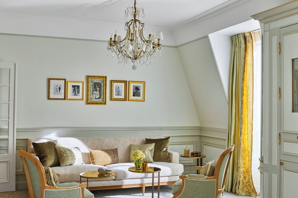A room with a sofa, two armchairs, a small round table, and a chandelier. There are four framed pictures on the wall.
