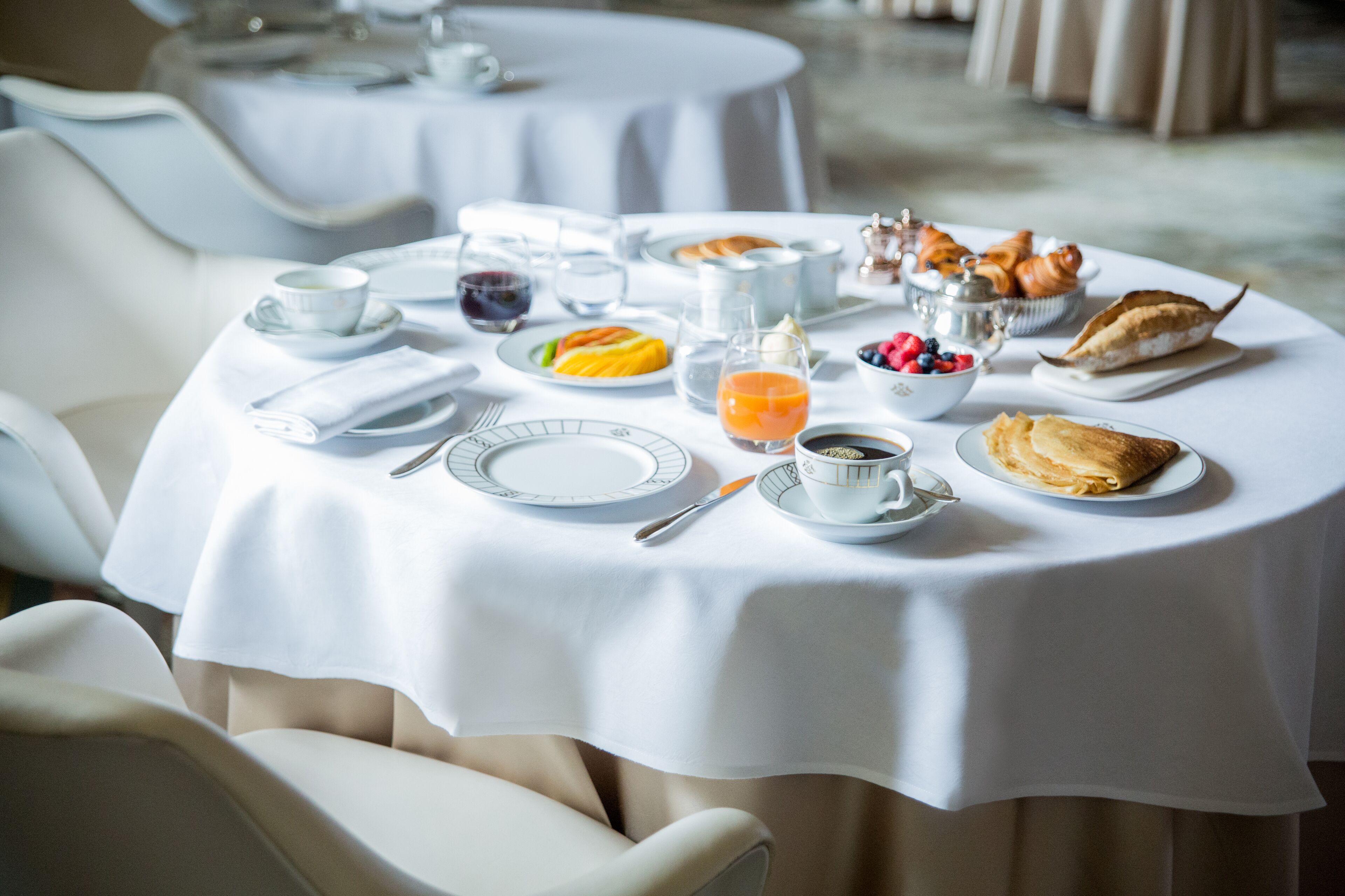 A breakfast setting with a white tablecloth, plates, cups, and a variety of food items including pastries and fruits.