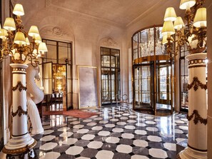 Un vestíbulo imponente con suelo a cuadros blanco y negro, columnas ornamentadas y una lámpara de araña grande.
