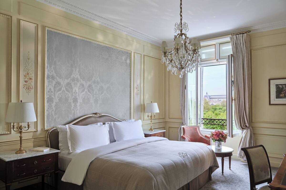 A hotel room with a large bed, two bedside tables with lamps, a chandelier, a window with curtains, and a view of greenery outside.