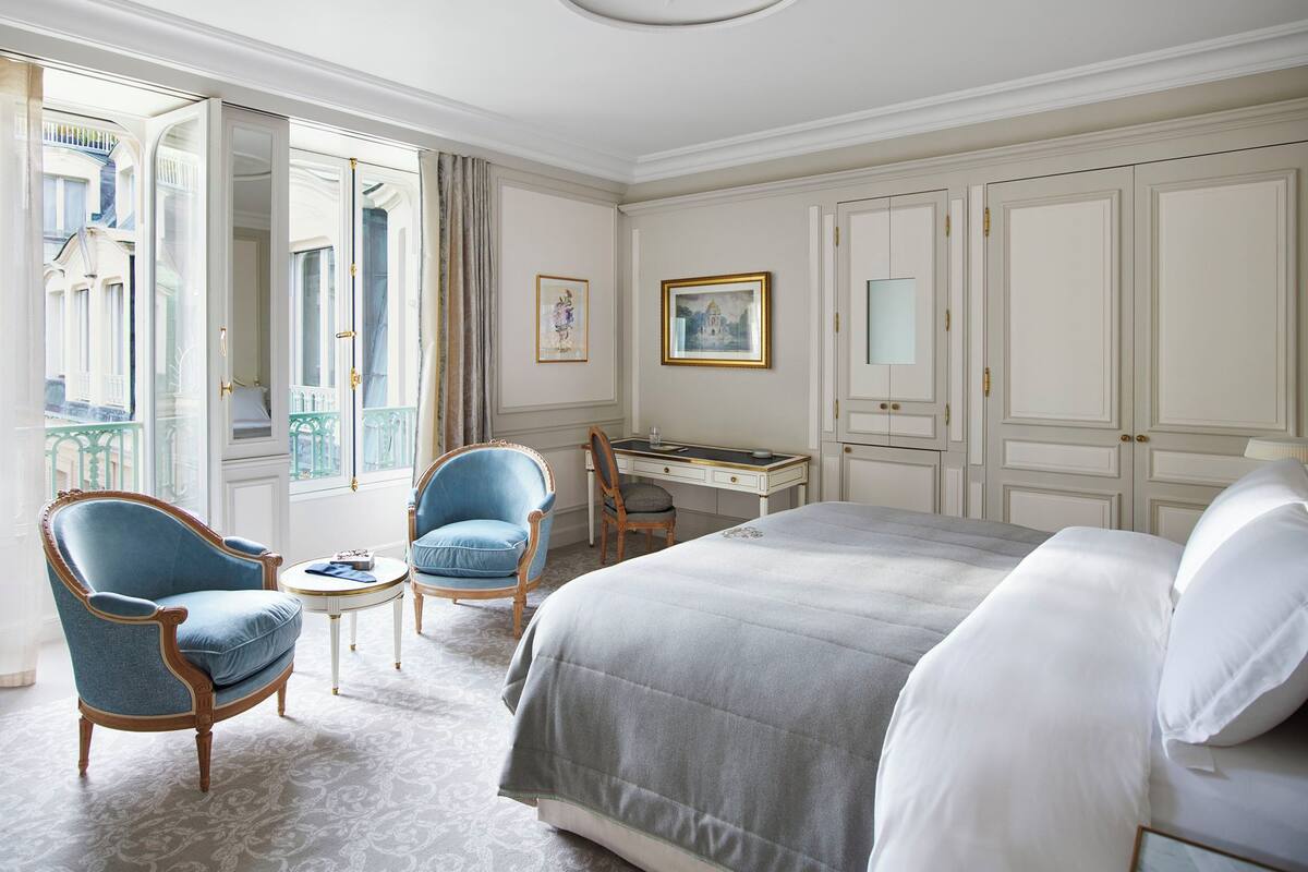 A hotel room with a bed, two armchairs, a desk, and a view of the outside.