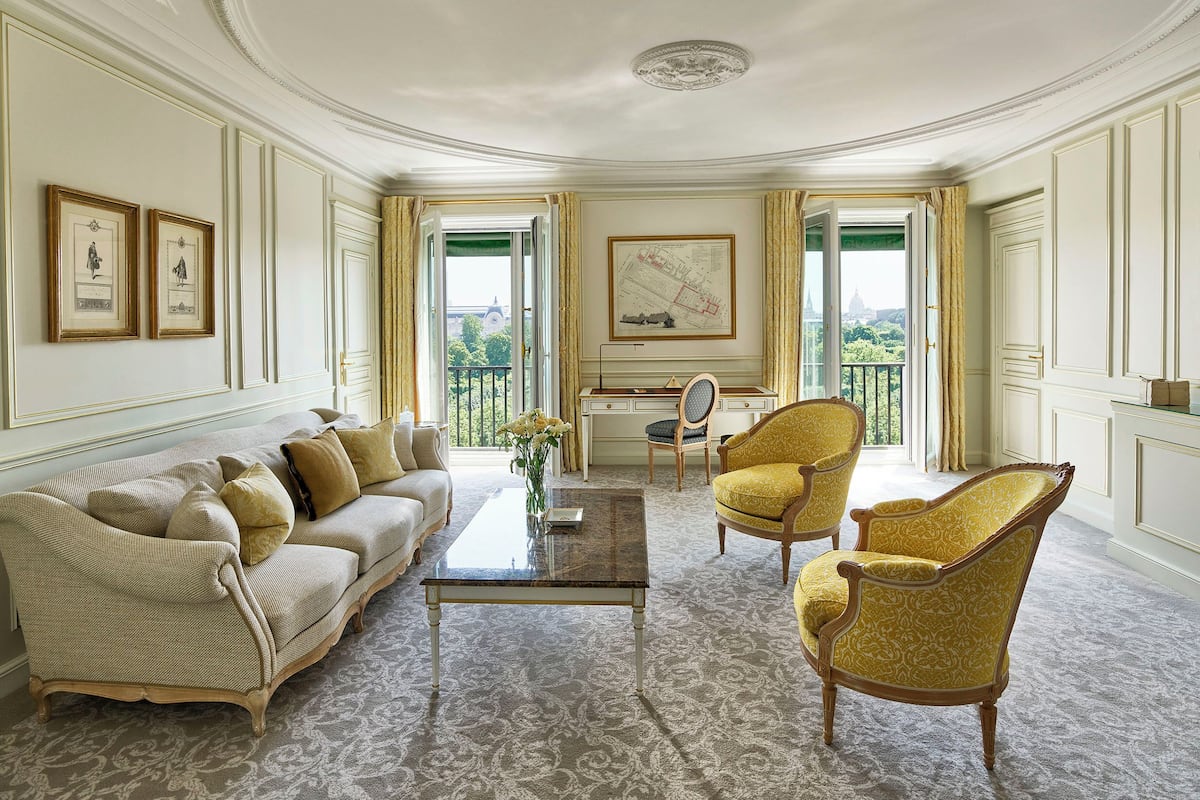A room with a sofa, two armchairs, a coffee table, and a desk with a chair. There are framed pictures on the wall, a map, and a view through a window.