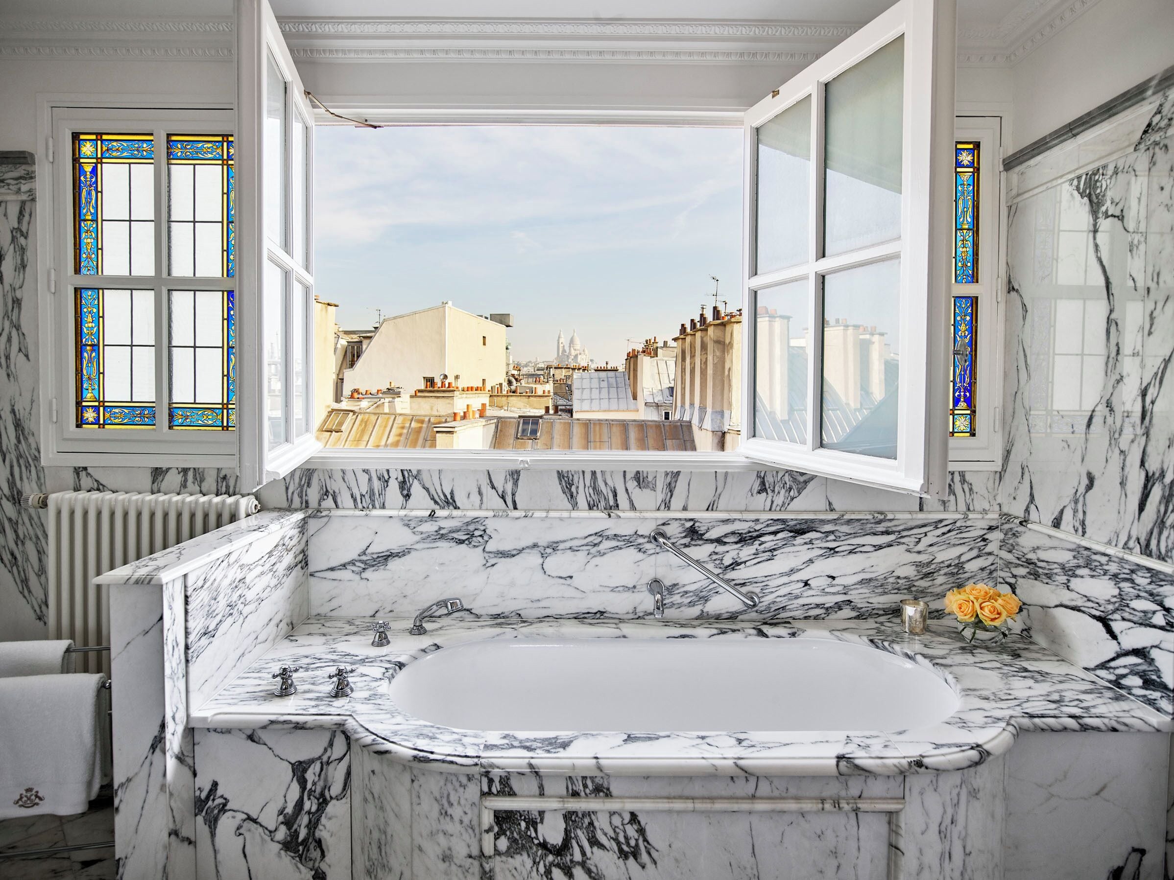 A bathroom with a large marble bathtub, a view of the cityscape through large windows, and a radiator.