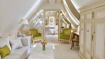 Een ruime kamer met een hoge plafond, een groot bed, een zithoek met een bank en stoelen, en uitzicht op een andere kamer door een groot raam.