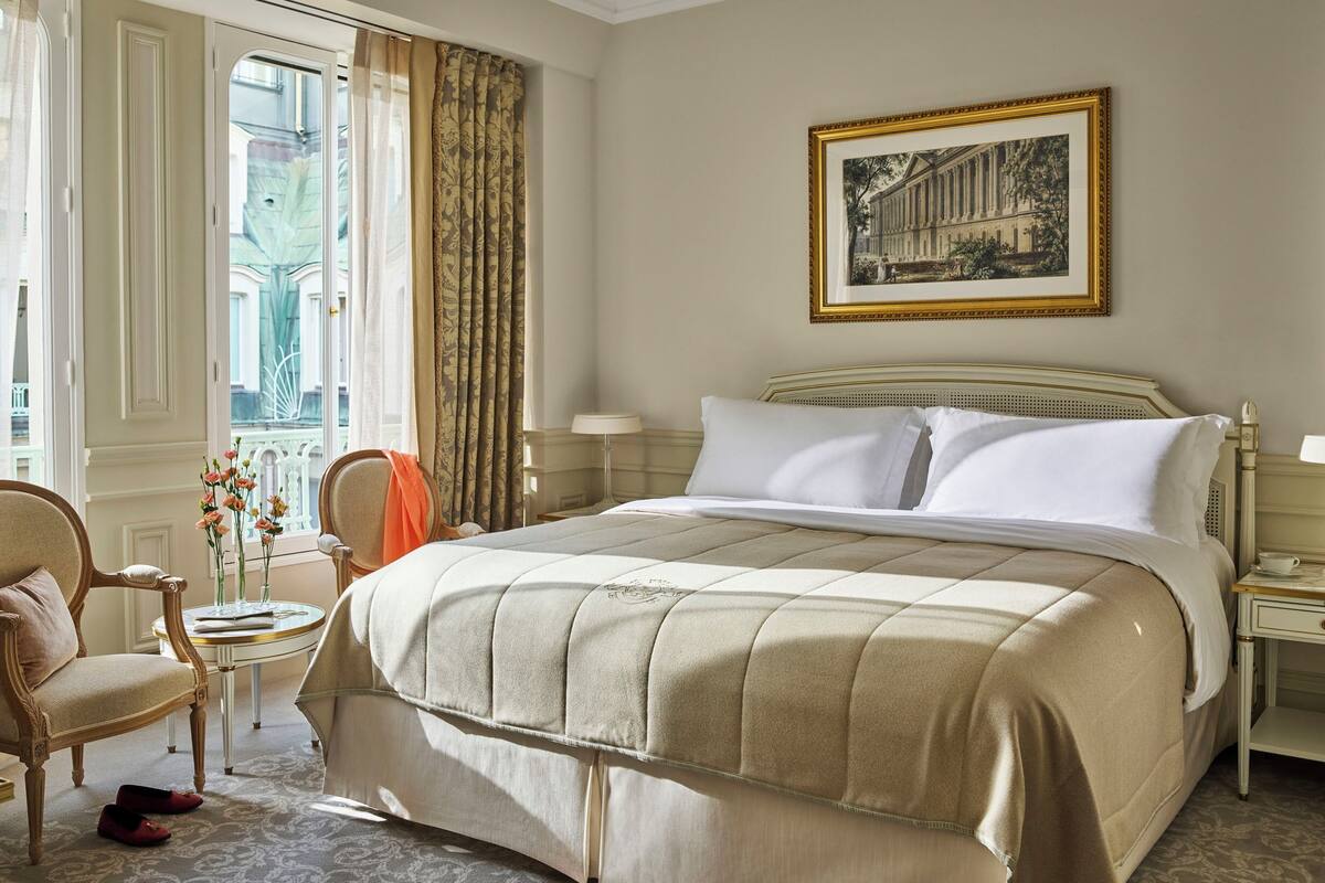 A hotel room with a large bed, a chair, a table with a vase of flowers, and a framed picture on the wall.