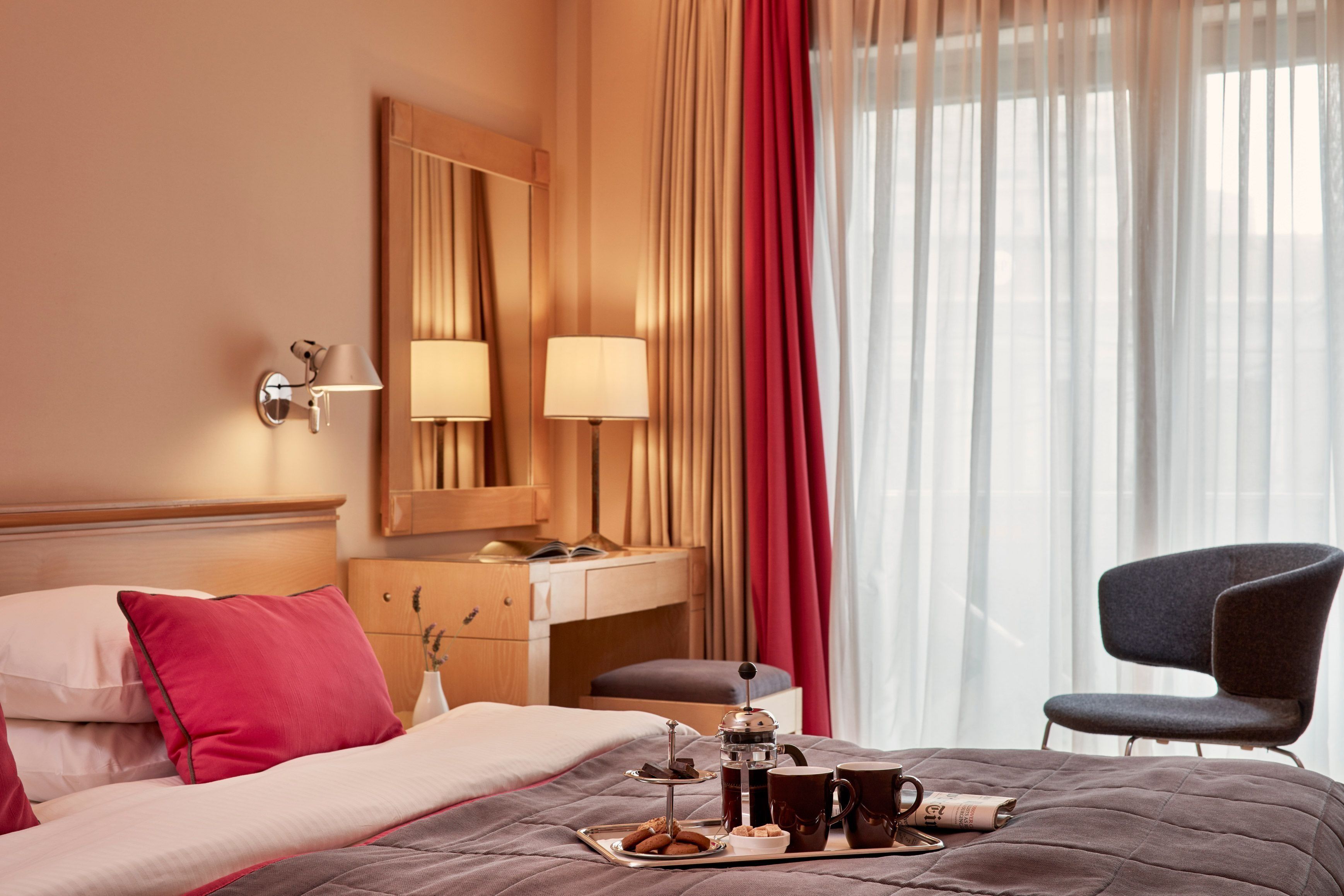 A hotel room with a bed, a desk, a chair, and a breakfast tray with pastries and coffee.
