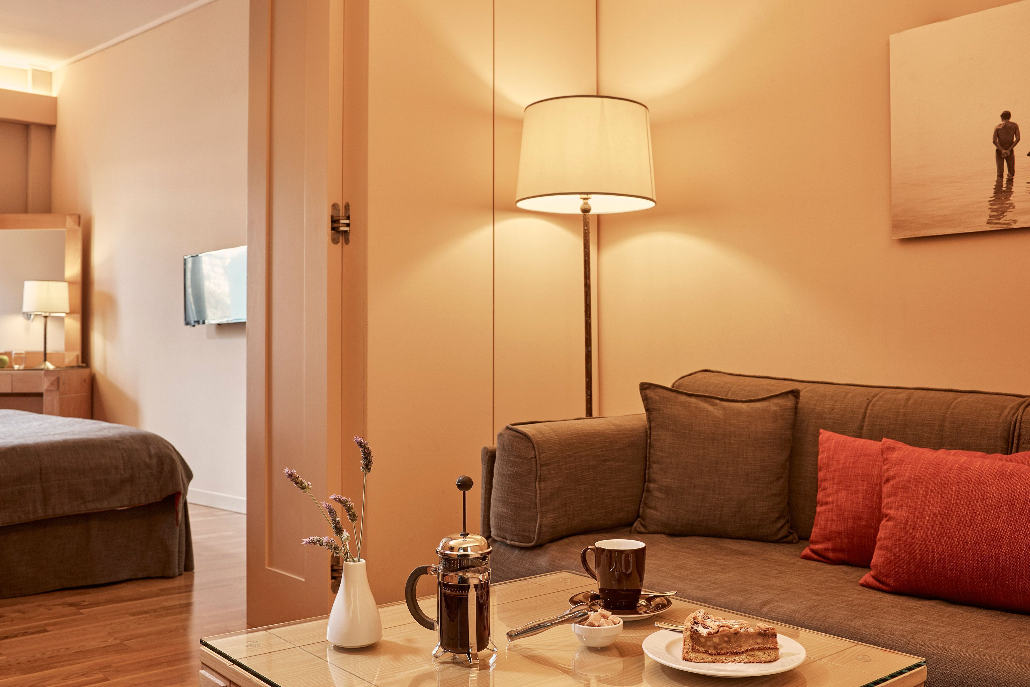 Una habitación de hotel acogedora con una cama, un sofá, una mesa de centro con una cafetera, una taza y una rebanada de pastel.