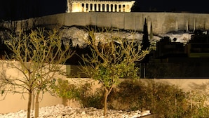 A nighttime view of a fountain with water jets, surrounded by a landscaped area with trees and a stone wall.