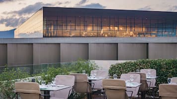 An outdoor dining area with tables and chairs, a modern building with large windows in the background, and a view of the sky.