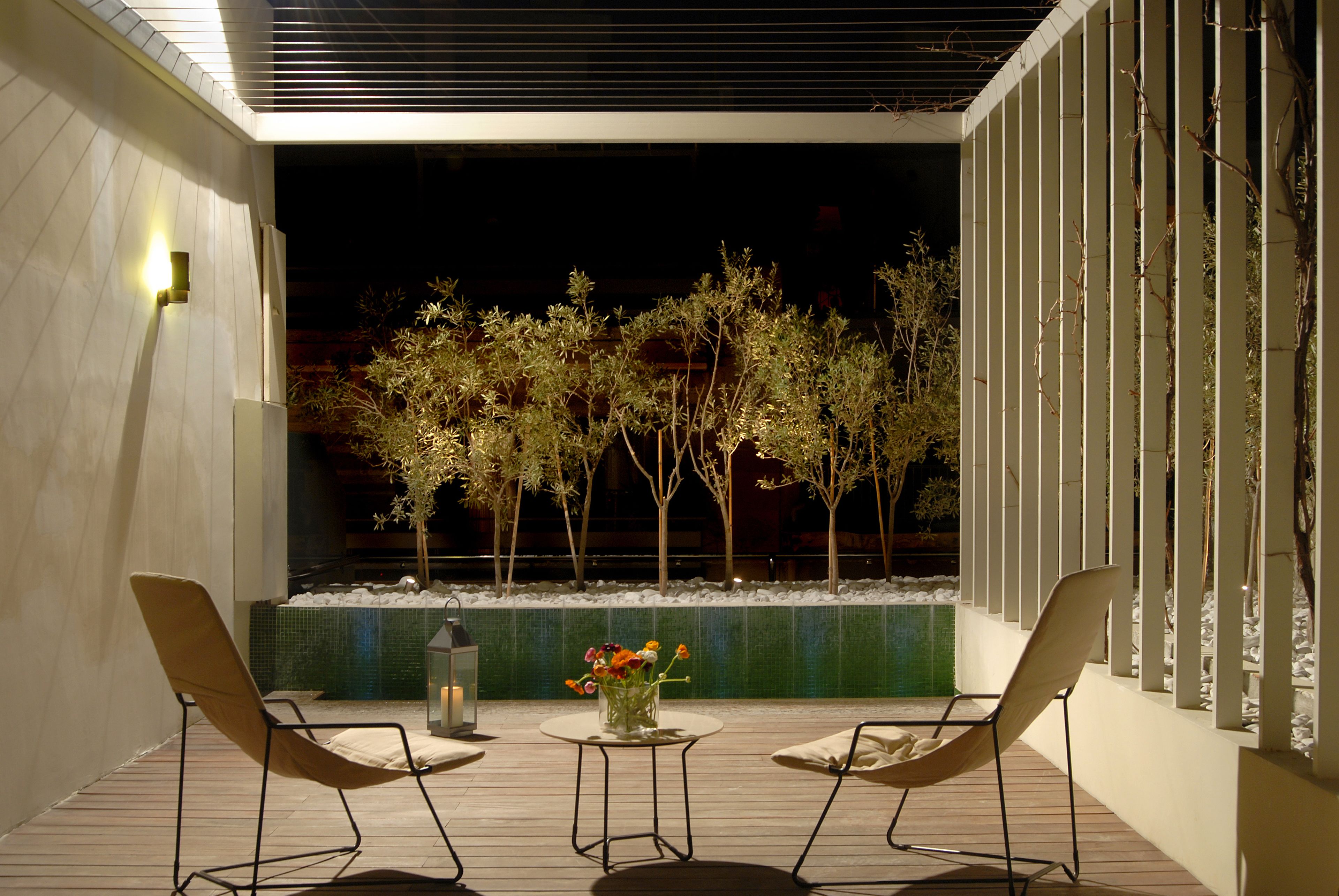 An outdoor seating area with two chairs and a table, a small vase with flowers, and a tiled wall with trees in the background.