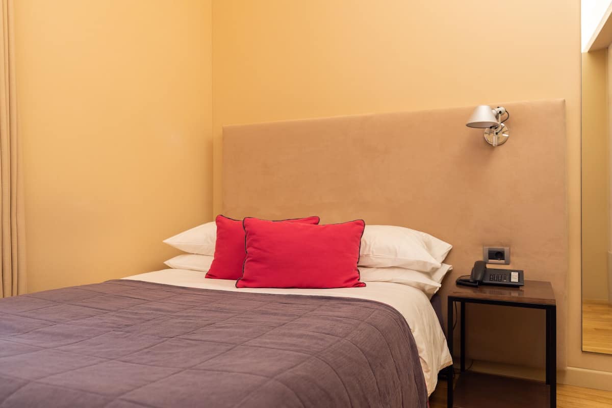 Un lit bien fait, avec une couvre-lit violette et deux oreillers rouges, une table de chevet avec une lampe et une petite enceinte.