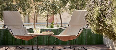 Two cushioned chairs with a small table between them, set on a wooden deck with a tiled wall and greenery in the background.