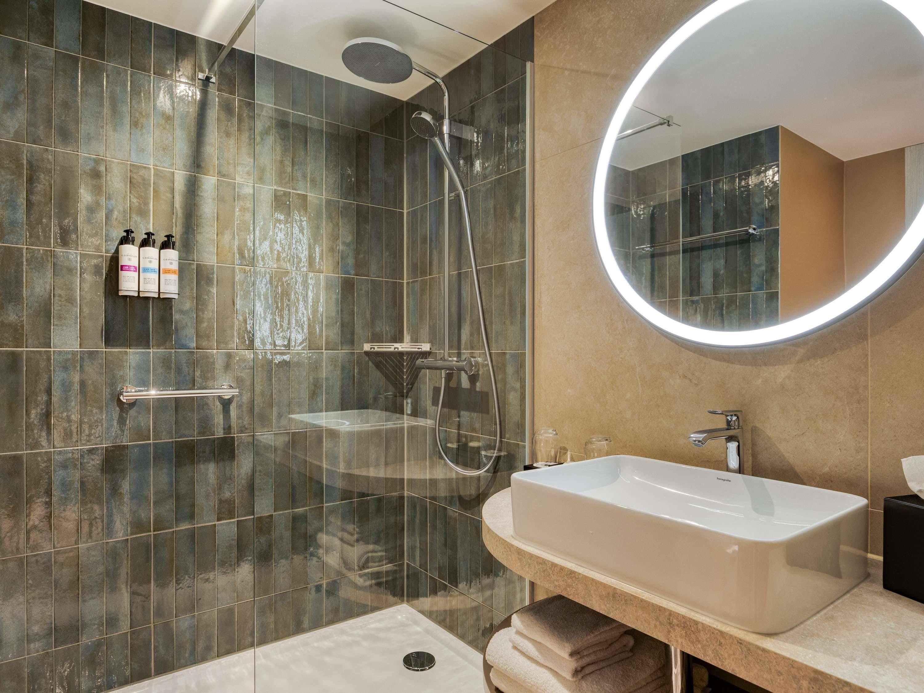 A modern bathroom with a glass-enclosed shower, a vanity with a sink, and a round mirror with a light.