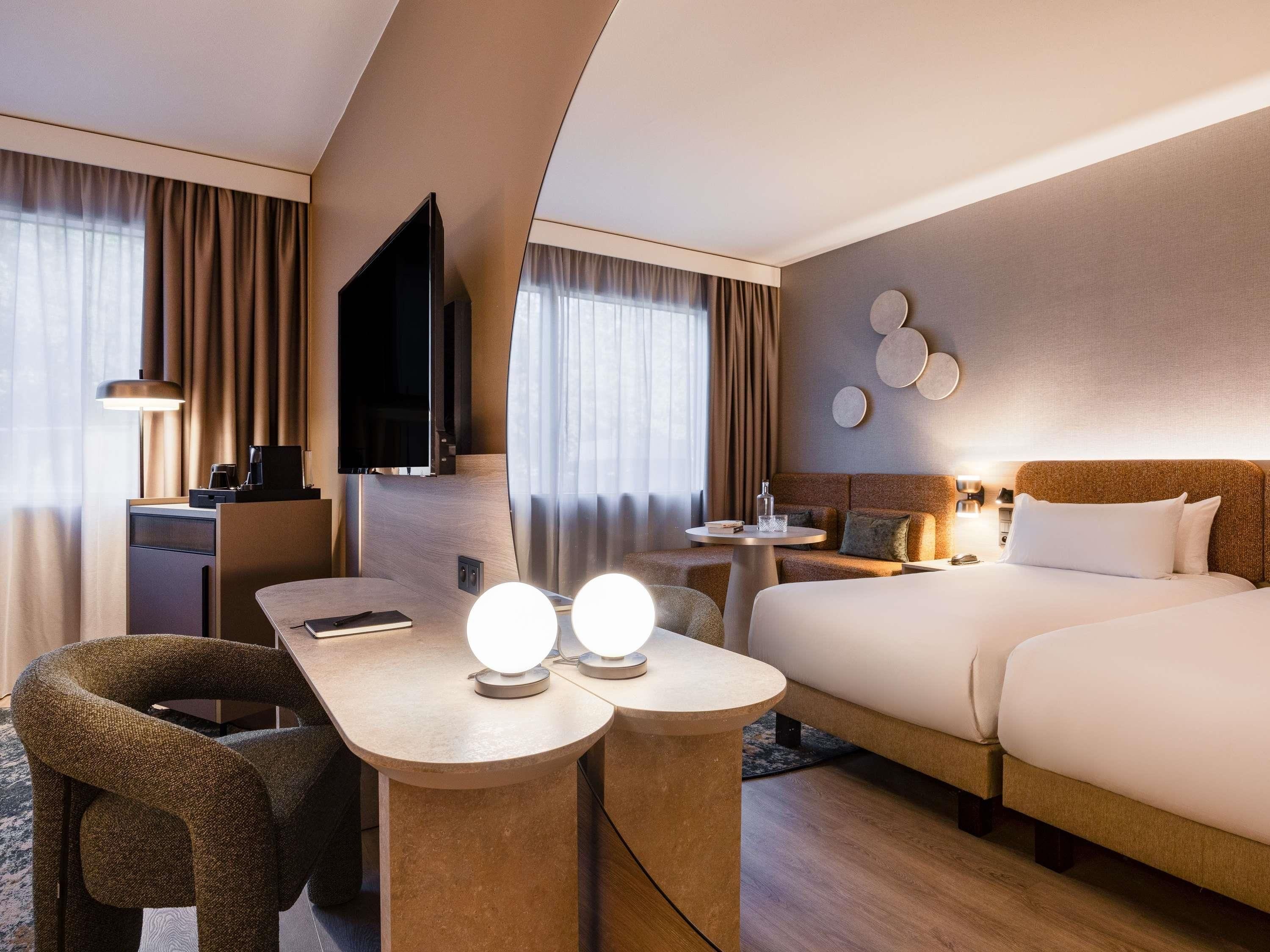 A hotel room with a bed, a desk, a chair, a TV, and a wall-mounted light fixture.