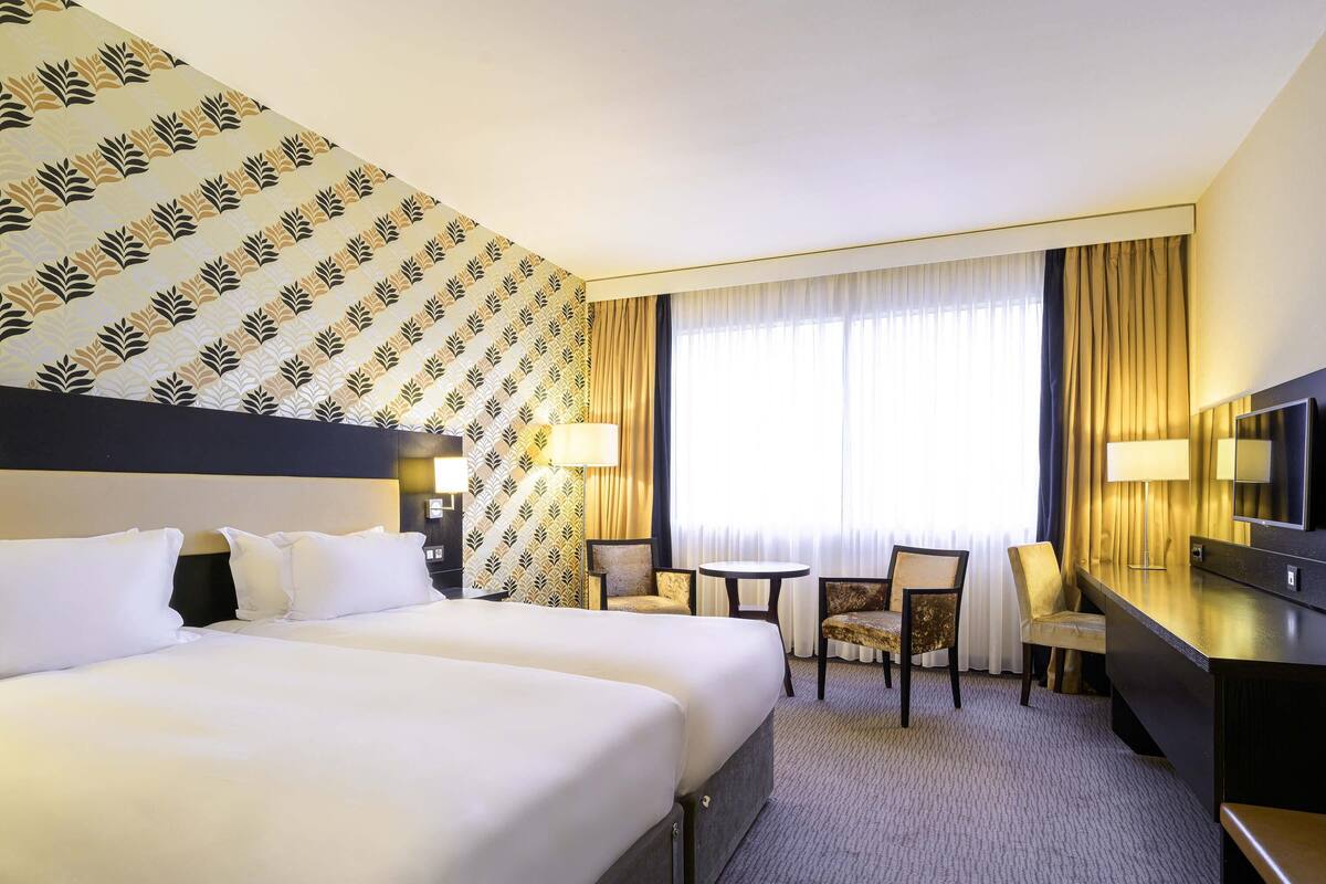 Habitación de hotel con una cama grande, un escritorio con silla, televisión y ventana con cortinas.