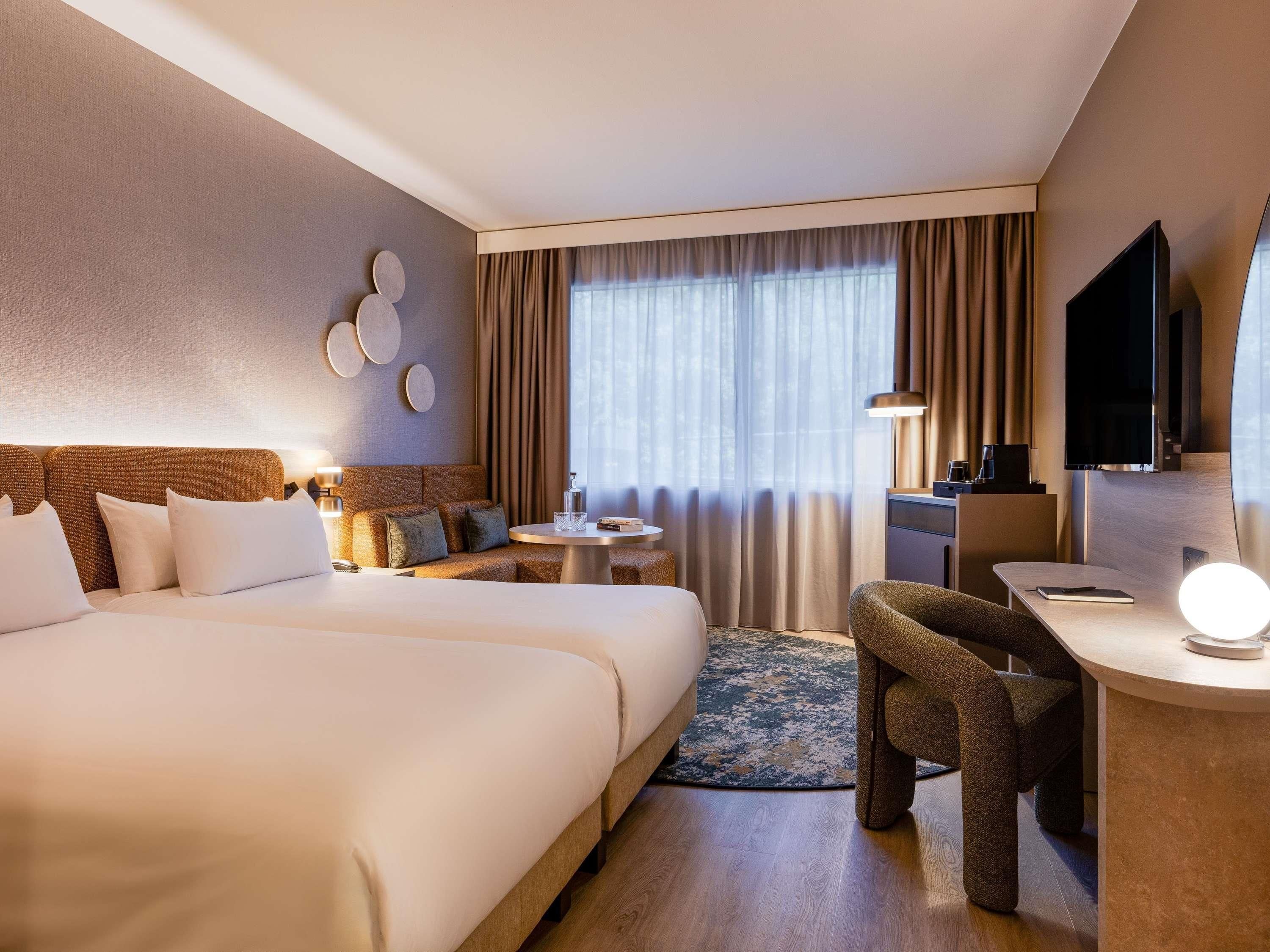 Una habitación de hotel con una cama grande, un escritorio con silla, un televisor y un ventanal con cortinas.