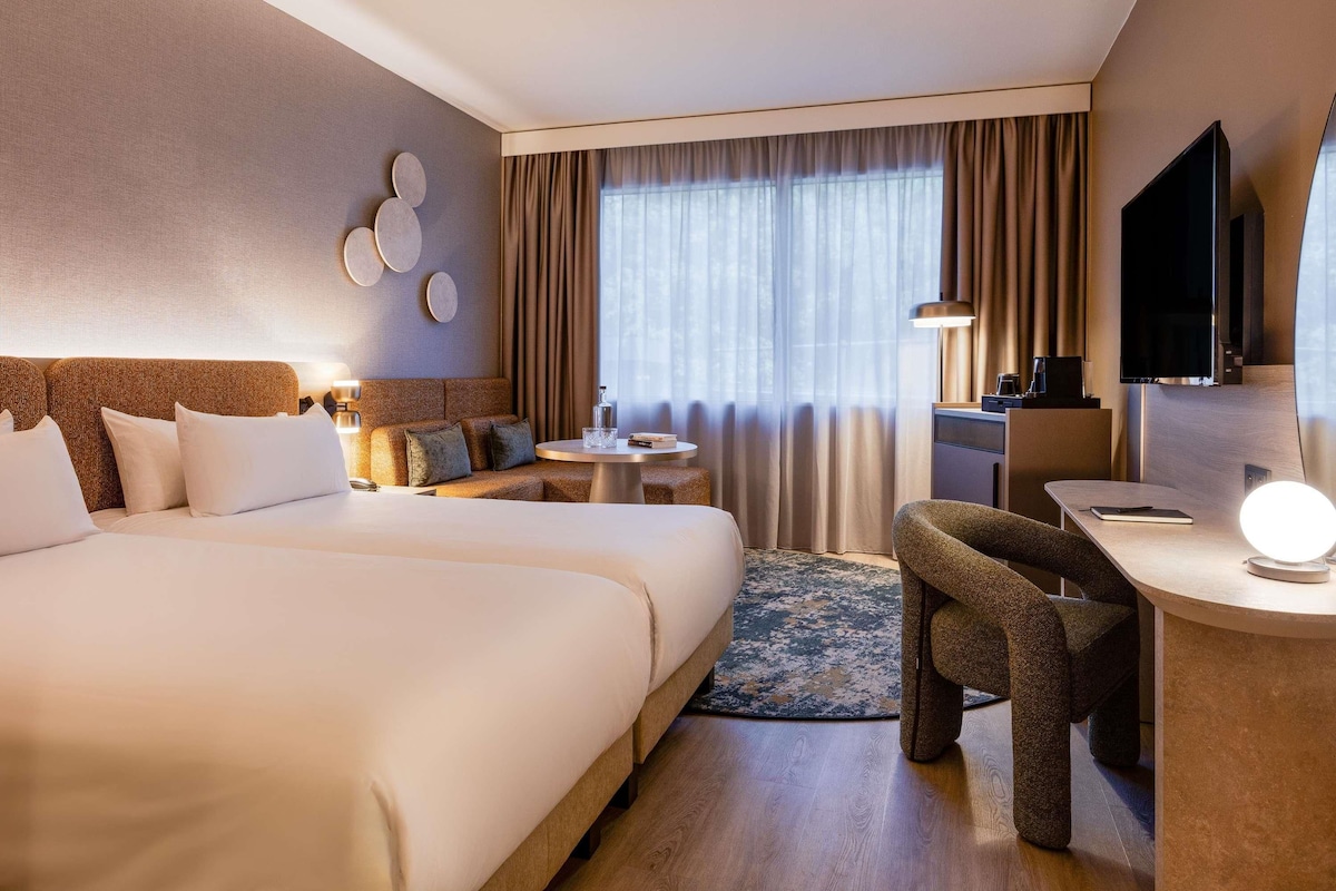 A hotel room with a large bed, a desk with a chair, a TV, and a window with curtains.