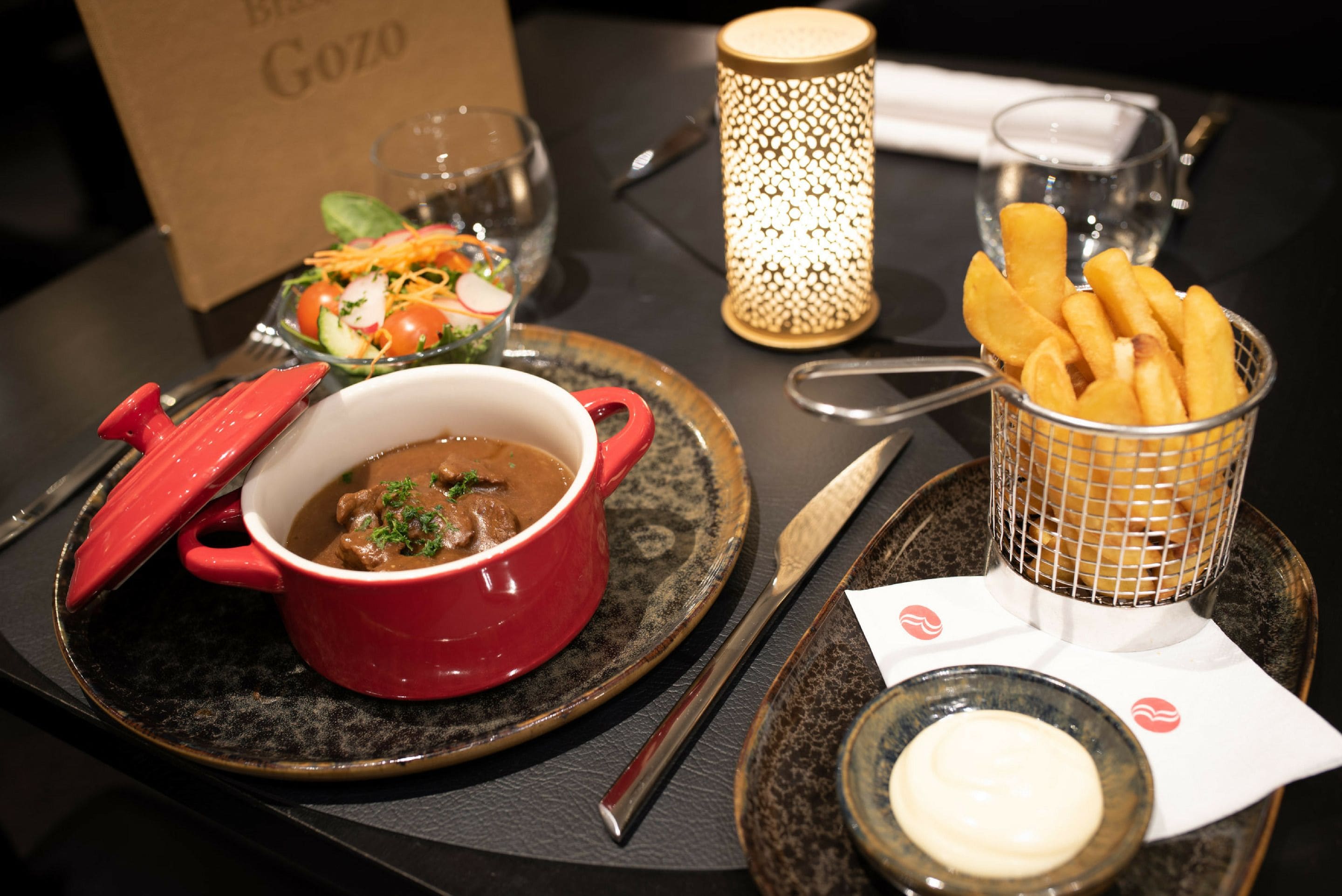 Una mesa de comedor con un tazón rojo de estofado, una ensalada pequeña, una cesta de papas fritas y una vela.