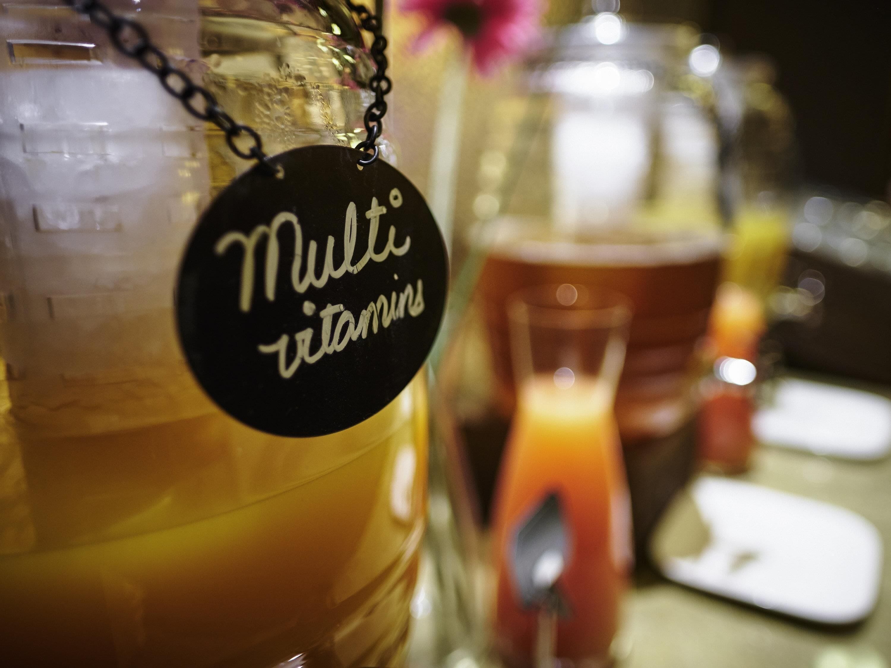 A close-up of a beverage dispenser labeled "Multi vitamins" with a glass of orange juice in the background.