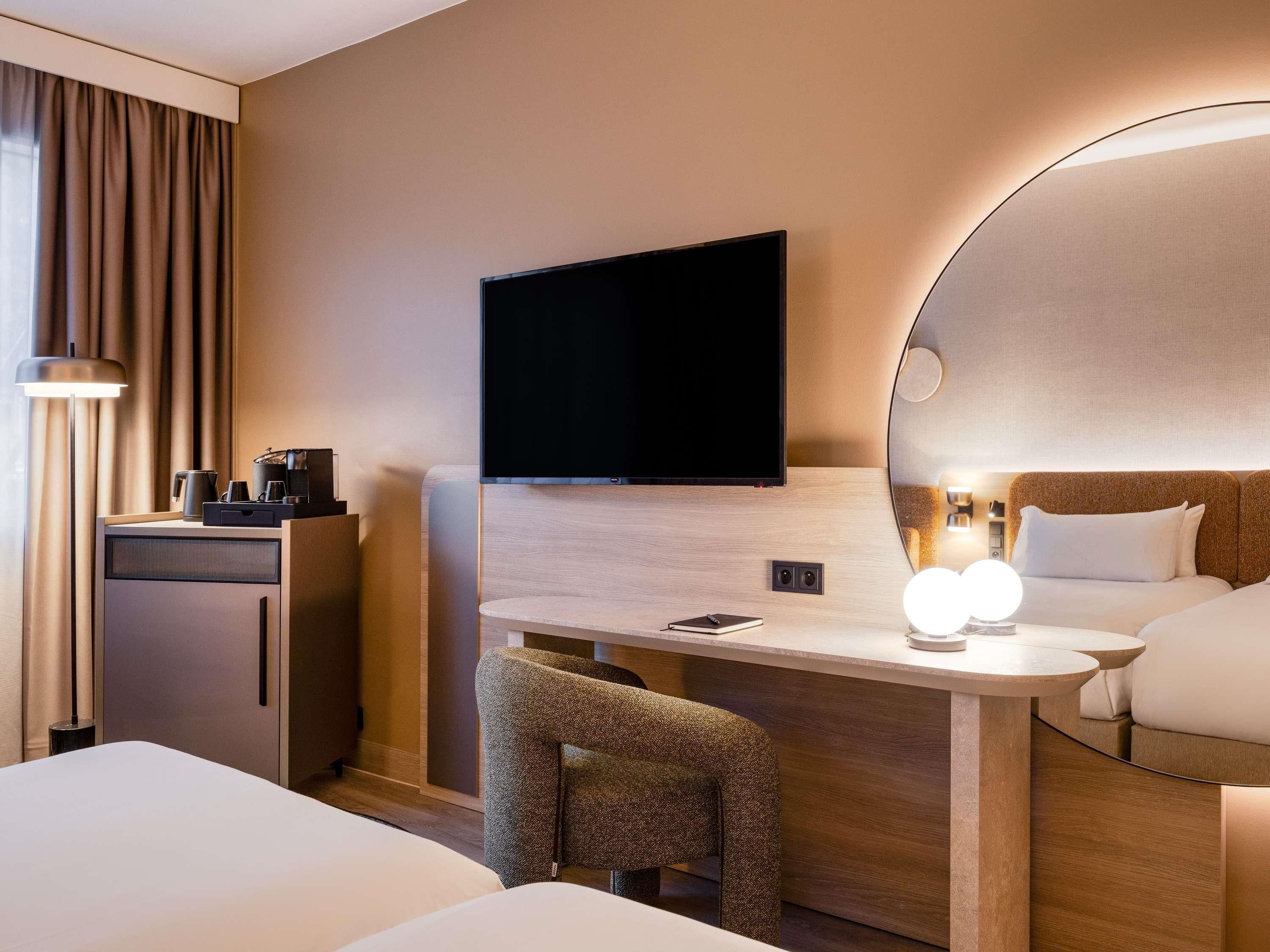 A hotel room with a bed, a desk with a flat-screen TV, a chair, a bedside table with a lamp, and a large mirror.