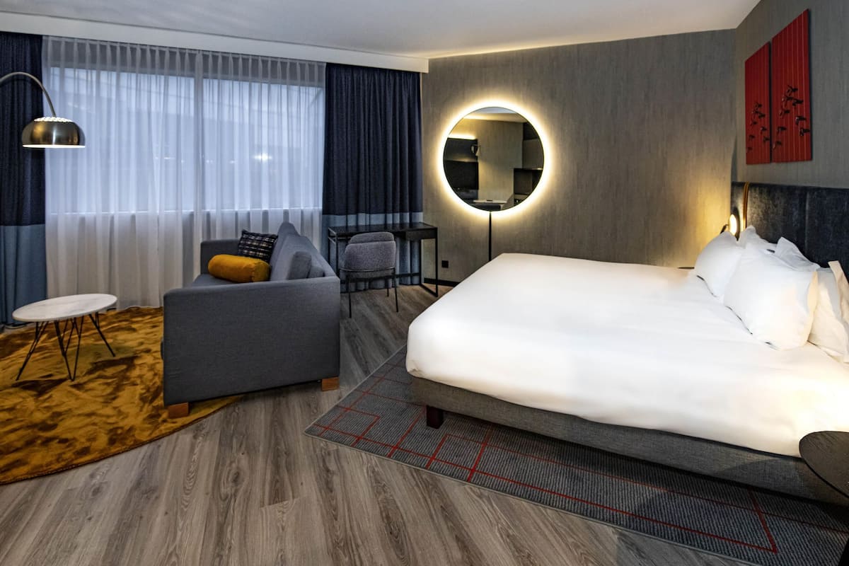 Una habitación de hotel moderna con una cama grande, un sofá, una mesita, una silla, un espejo redondo y un escritorio.