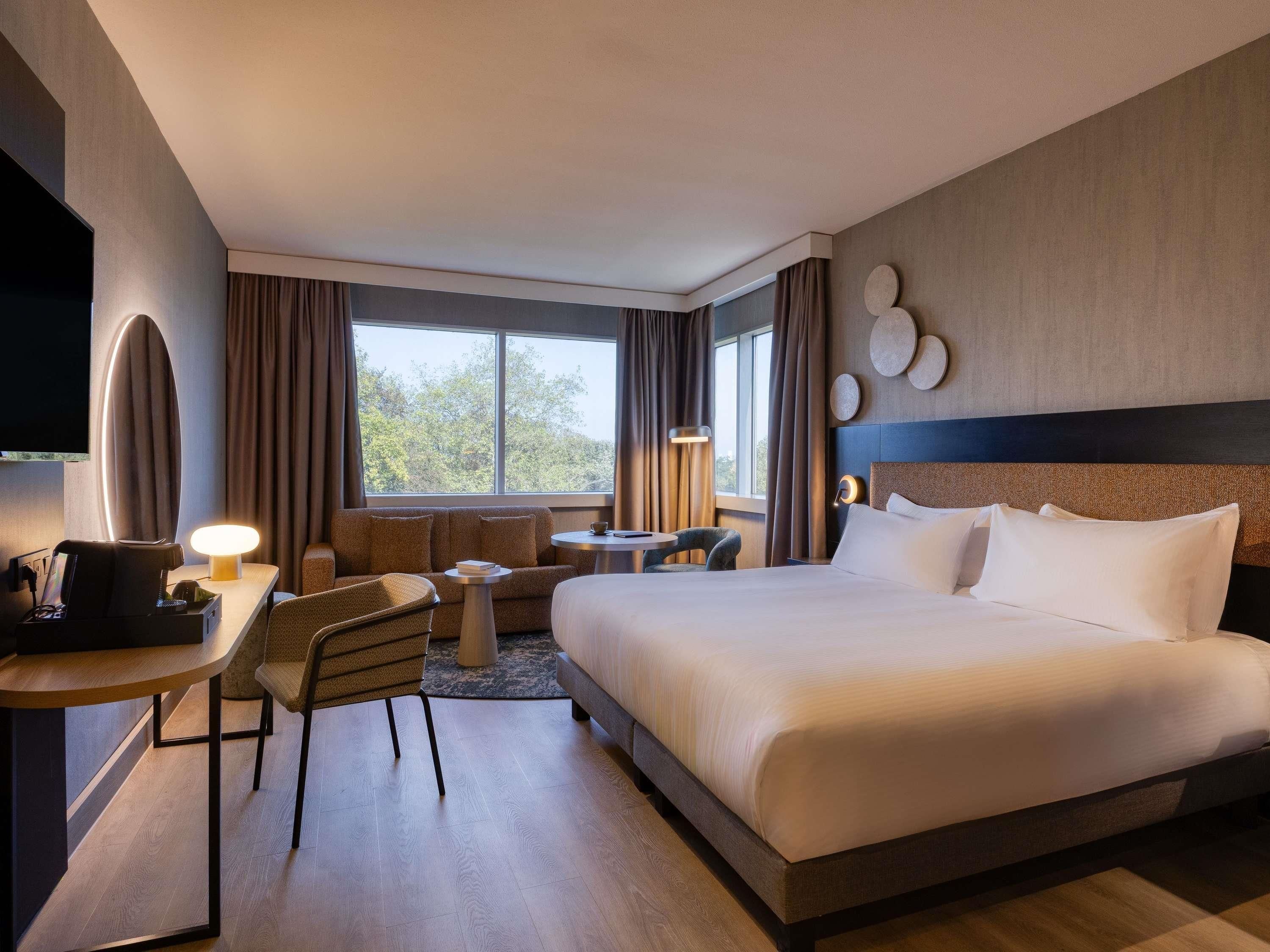 A hotel room with a bed, a desk with a chair, a TV, and a window with curtains.