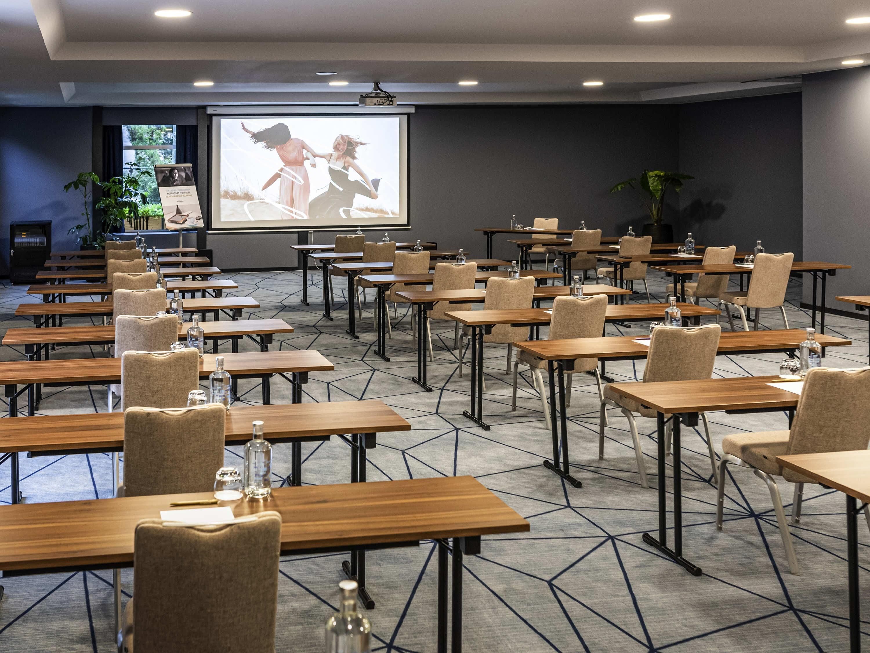A conference room with rows of tables and chairs, a projector screen, and a presentation on the wall.
