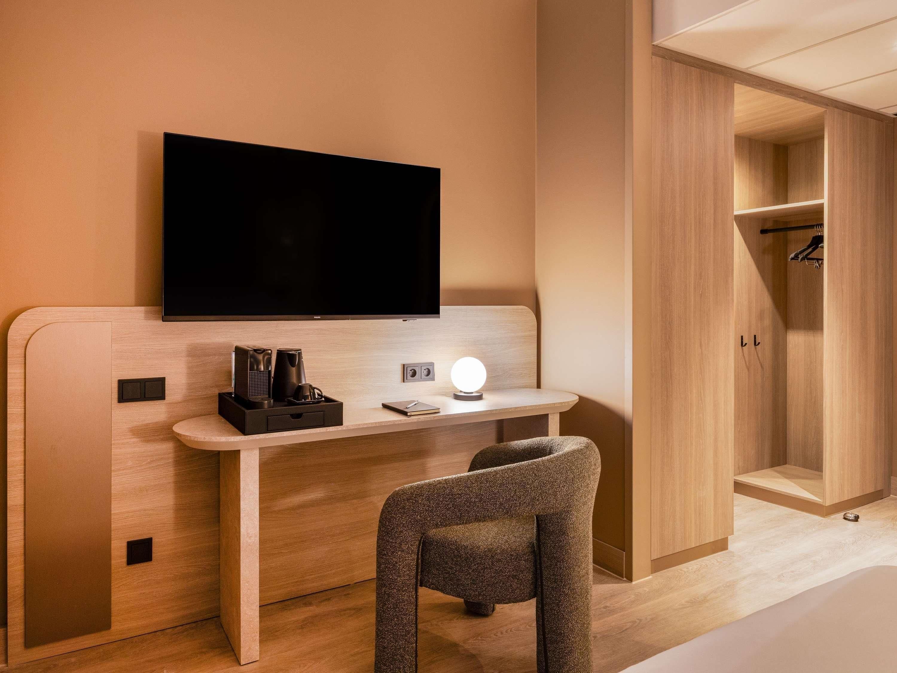 A hotel room with a flat-screen TV mounted on a wall, a bedside table with a lamp, a chair, and a wardrobe with a mirror.