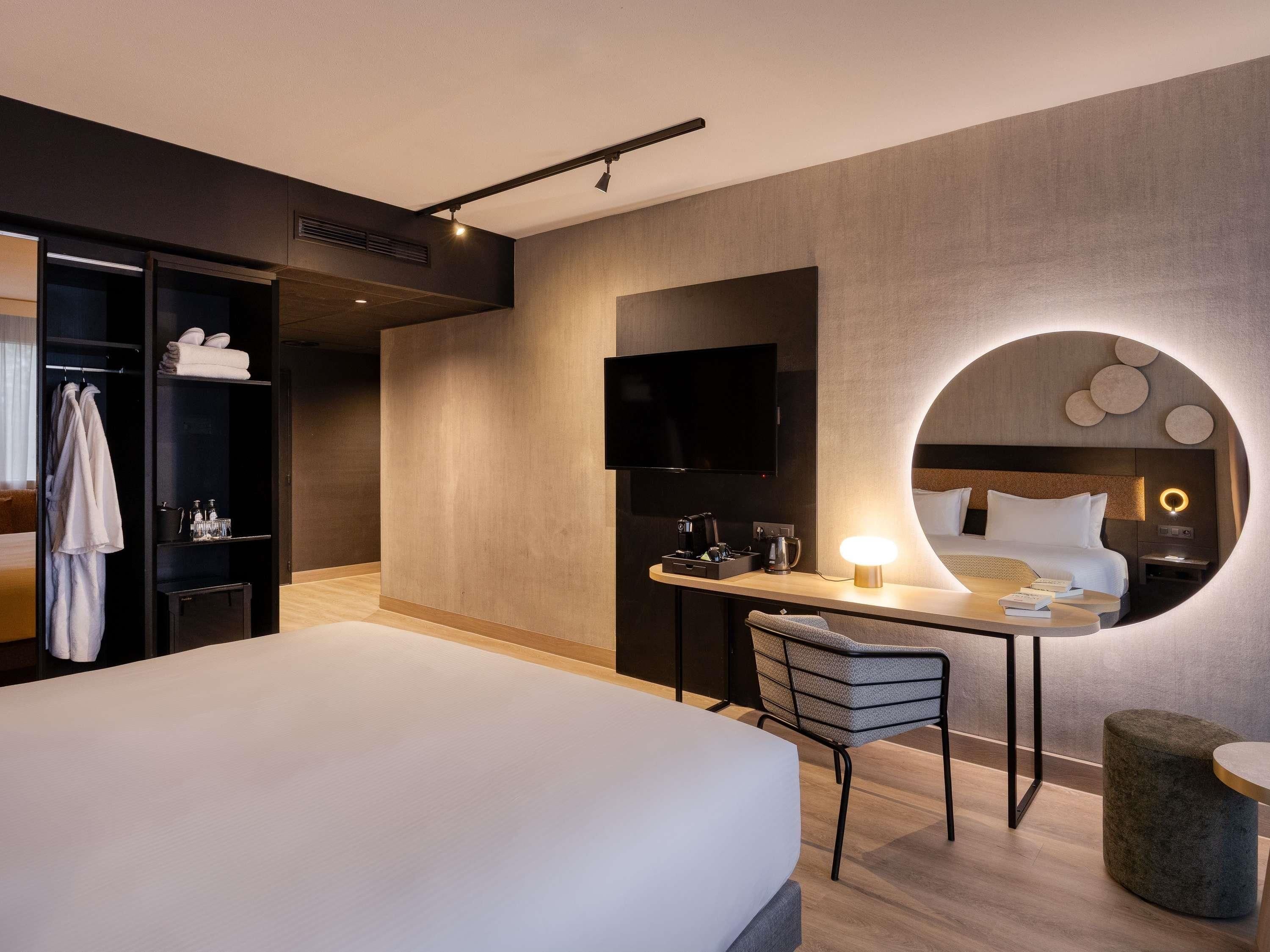Une chambre d’hôtel moderne équipée d’un lit, d’un bureau, d’une chaise, d’une télévision et d’un miroir rond.